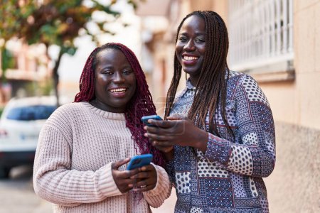 Téléchargez les photos : Afro-américaine femmes amis souriant confiant en utilisant smartphone à la rue - en image libre de droit