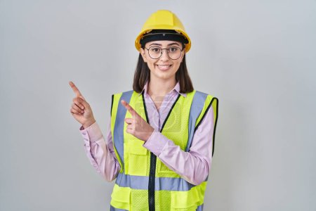 Foto de Chica hispana vistiendo uniforme de constructor y hardhat sonriendo y mirando a la cámara apuntando con dos manos y dedos hacia un lado. - Imagen libre de derechos