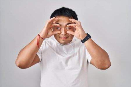 Téléchargez les photos : Young chinese man standing over white background trying to open eyes with fingers, sleepy and tired for morning fatigue - en image libre de droit