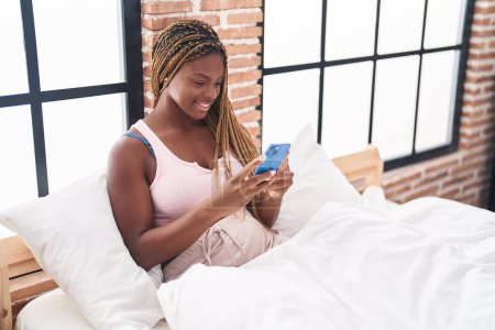 Foto de Mujer afroamericana usando teléfono inteligente sentado en la cama en el dormitorio - Imagen libre de derechos