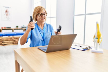 Photo for Middle age blonde woman wearing physio therapy uniform doing tele rehab using hand grip at clinic - Royalty Free Image