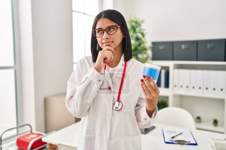 Photo for Young hispanic doctor woman holding cotton buds serious face thinking about question with hand on chin, thoughtful about confusing idea - Royalty Free Image