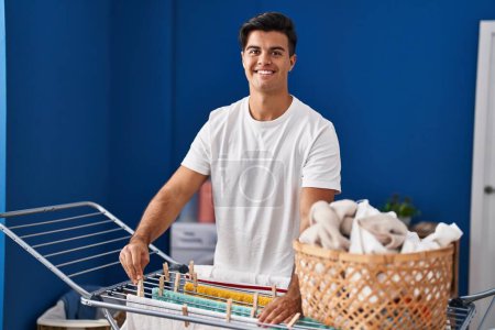 Foto de Joven hombre hispano sonriendo confiado colgando ropa en tendedero en la lavandería - Imagen libre de derechos