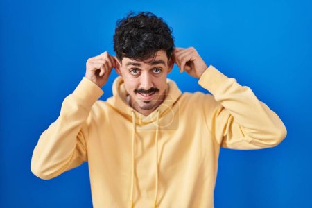 Photo for Hispanic man standing over blue background smiling pulling ears with fingers, funny gesture. audition problem - Royalty Free Image