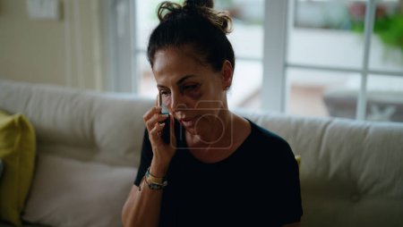 Foto de Mujer hispana de mediana edad que sufre por violencia doméstica hablando en el teléfono inteligente en casa - Imagen libre de derechos