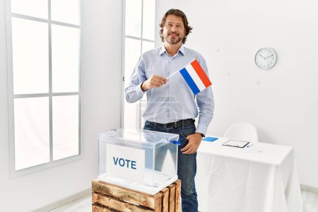 Foto de Hombre caucásico de mediana edad sonriendo confiado sosteniendo la bandera holandesa votando en colegio electoral - Imagen libre de derechos