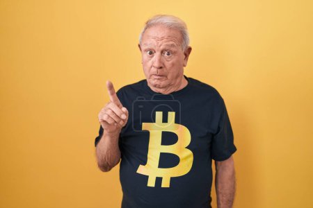 Foto de Hombre mayor con el pelo gris usando camiseta bitcoin señalando a un lado preocupado y nervioso con el dedo índice, preocupado y expresión sorprendida - Imagen libre de derechos
