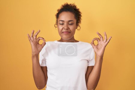 Photo for Young hispanic woman with curly hair standing over yellow background relaxed and smiling with eyes closed doing meditation gesture with fingers. yoga concept. - Royalty Free Image