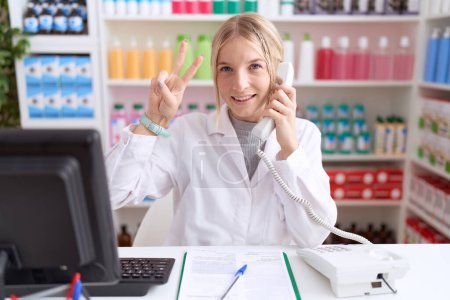Téléchargez les photos : Jeune femme caucasienne travaillant à la pharmacie pharmacie parlant au téléphone souriant en regardant la caméra montrant des doigts faisant signe de victoire. numéro deux. - en image libre de droit