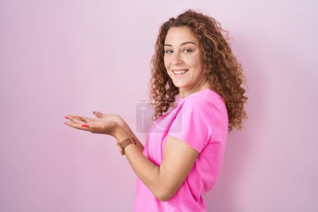 Foto de Joven mujer caucásica de pie sobre fondo rosa señalando a un lado con las manos abiertas palmeras mostrando espacio de copia, presentando anuncio sonriente emocionado feliz - Imagen libre de derechos