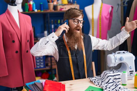 Téléchargez les photos : Young redhead man tailor talking on smartphone with unhappy expression at clothing factory - en image libre de droit