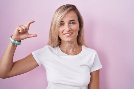 Téléchargez les photos : Jeune femme blonde debout sur fond rose geste souriant et confiant avec la main faisant signe de petite taille avec les doigts regardant et la caméra. concept de mesure. - en image libre de droit