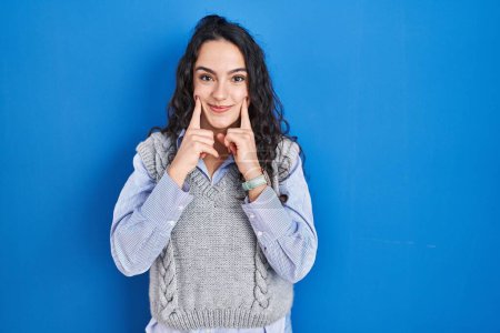 Téléchargez les photos : Jeune femme brune debout sur fond bleu souriant avec la bouche ouverte, les doigts pointant et forçant sourire joyeux - en image libre de droit