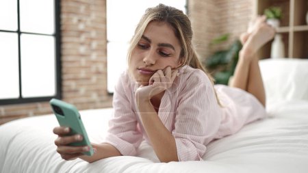 Téléchargez les photos : Jeune femme blonde utilisant un smartphone avec une expression triste dans la chambre - en image libre de droit