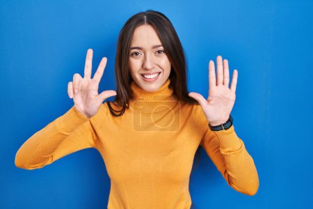 Photo for Young brunette woman standing over blue background showing and pointing up with fingers number eight while smiling confident and happy. - Royalty Free Image
