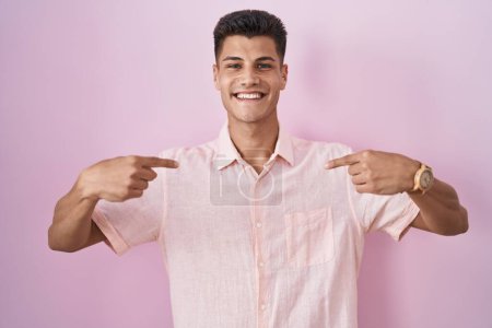 Foto de Joven hombre hispano de pie sobre fondo rosa mirando confiado con sonrisa en la cara, señalándose con los dedos orgullosos y felices. - Imagen libre de derechos