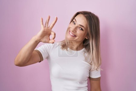 Photo for Blonde caucasian woman standing over pink background smiling positive doing ok sign with hand and fingers. successful expression. - Royalty Free Image