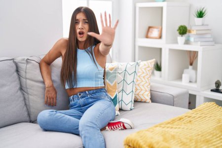 Photo for Young brunette woman sitting on the sofa at home doing stop gesture with hands palms, angry and frustration expression - Royalty Free Image
