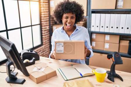 Photo for Black woman with curly hair working at small business ecommerce holding box winking looking at the camera with sexy expression, cheerful and happy face. - Royalty Free Image