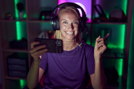 Téléchargez les photos : Young gamer woman playing video games with smartphone smiling happy pointing with hand and finger to the side - en image libre de droit