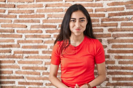 Foto de Young teenager girl standing over bricks wall puffing cheeks with funny face. mouth inflated with air, crazy expression. - Imagen libre de derechos