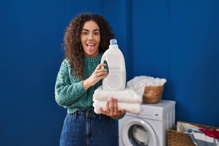 Foto de Young hispanic woman holding laundry and detergent bottle sticking tongue out happy with funny expression. - Imagen libre de derechos