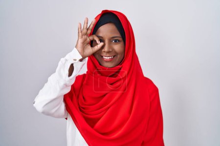 Photo for Young arab woman wearing traditional islamic hijab scarf doing ok gesture with hand smiling, eye looking through fingers with happy face. - Royalty Free Image