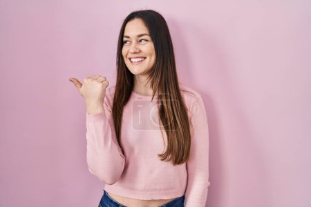 Téléchargez les photos : Jeune femme brune debout sur fond rose souriant avec le visage heureux regardant et pointant vers le côté avec le pouce vers le haut. - en image libre de droit