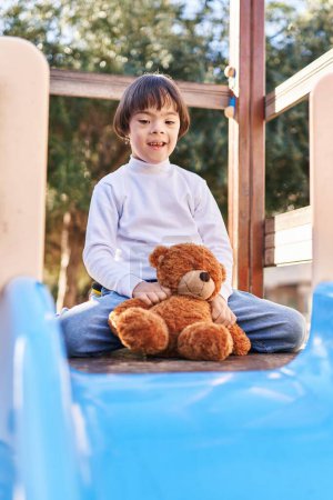 Foto de Síndrome de Down niño sonriendo confiado jugando con osito de peluche en la diapositiva en el parque - Imagen libre de derechos