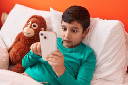 Photo for Adorable hispanic boy using smartphone lying on bed with monkey doll at bedroom - Royalty Free Image