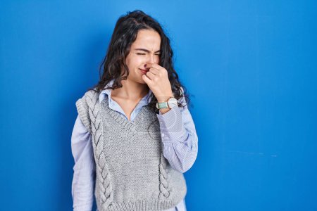 Téléchargez les photos : Jeune femme brune debout sur fond bleu sentant quelque chose de puant et dégoûtant, odeur intolérable, retenant la respiration avec les doigts sur le nez. mauvaise odeur - en image libre de droit