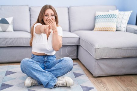 Foto de Young caucasian woman sitting on the floor at the living room laughing at you, pointing finger to the camera with hand over mouth, shame expression - Imagen libre de derechos