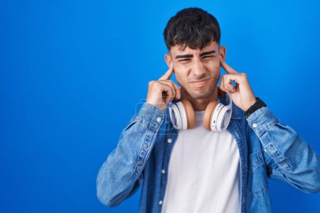 Foto de Joven hispano de pie sobre fondo azul cubriendo las orejas con los dedos con expresión molesta por el ruido de la música alta. concepto de sordo. - Imagen libre de derechos