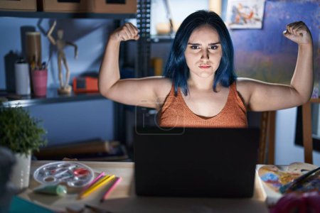 Photo for Young modern girl with blue hair sitting at art studio with laptop at night showing arms muscles smiling proud. fitness concept. - Royalty Free Image