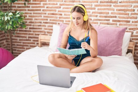 Téléchargez les photos : Jeune étudiante blonde écoutant de la musique étudier sur le lit dans la chambre - en image libre de droit