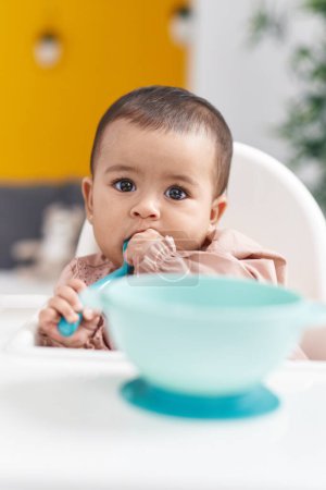 Photo for Adorable hispanic baby sitting on highchair sucking spoon at home - Royalty Free Image