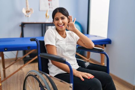 Téléchargez les photos : Young hispanic woman sitting on wheelchair at physiotherapy clinic smiling with hand over ear listening and hearing to rumor or gossip. deafness concept. - en image libre de droit