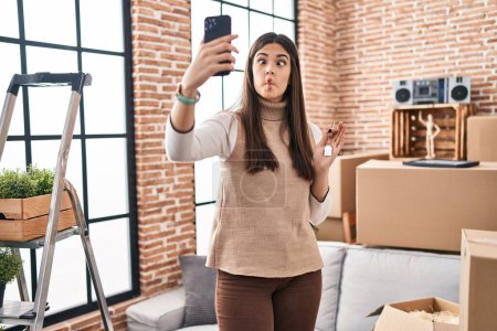 Photo for Young brunette woman moving to a new home doing video call making fish face with mouth and squinting eyes, crazy and comical. - Royalty Free Image