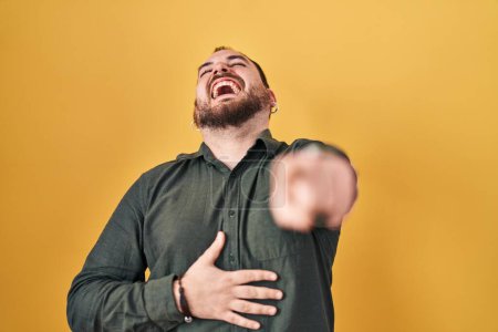 Photo for Plus size hispanic man with beard standing over yellow background laughing at you, pointing finger to the camera with hand over body, shame expression - Royalty Free Image