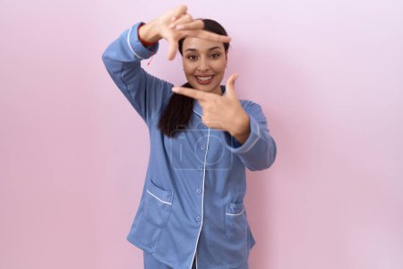 Téléchargez les photos : Jeune femme arabe portant un pyjama bleu souriant faisant cadre avec les mains et les doigts avec le visage heureux. concept de créativité et photographie. - en image libre de droit