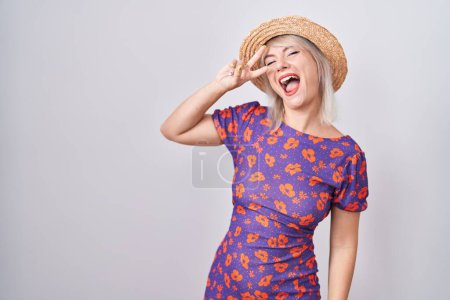Téléchargez les photos : Jeune femme caucasienne portant des fleurs robe et chapeau d'été faisant symbole de paix avec les doigts sur le visage, souriant joyeuse montrant la victoire - en image libre de droit