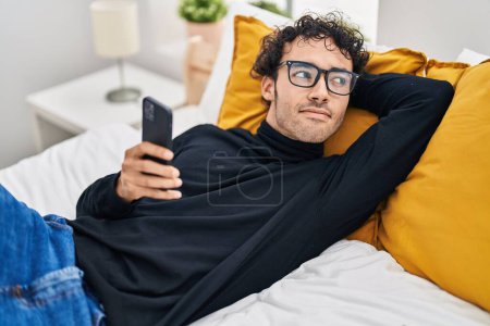 Photo pour Jeune homme hispanique utilisant smartphone couché sur le lit dans la chambre - image libre de droit