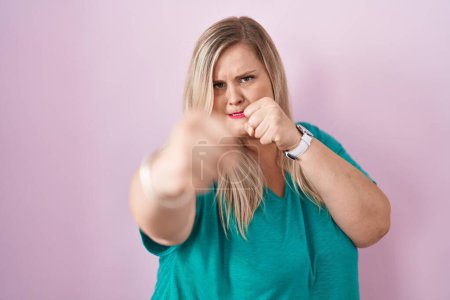 Téléchargez les photos : Caucasian plus size woman standing over pink background punching fist to fight, aggressive and angry attack, threat and violence - en image libre de droit