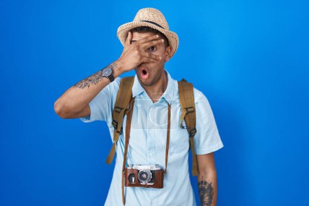 Téléchargez les photos : Jeune homme brésilien tenant caméra vintage regardant en état de choc couvrant le visage et les yeux avec la main, regardant à travers les doigts avec une expression embarrassée. - en image libre de droit