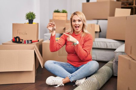 Téléchargez les photos : Blonde woman holding keys of new home sitting on the floor smiling happy pointing with hand and finger - en image libre de droit