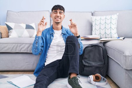 Photo for Young hispanic man sitting on the floor studying for university gesturing finger crossed smiling with hope and eyes closed. luck and superstitious concept. - Royalty Free Image