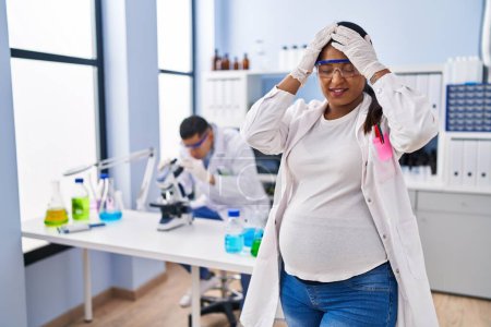 Foto de Mujer hispana joven esperando un bebé que trabaja en el laboratorio científico que sufre de dolor de cabeza desesperado y estresado debido al dolor y la migraña. manos en la cabeza. - Imagen libre de derechos