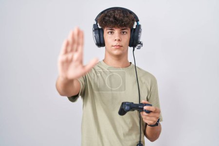 Foto de Hispanic teenager playing video game holding controller doing stop sing with palm of the hand. warning expression with negative and serious gesture on the face. - Imagen libre de derechos