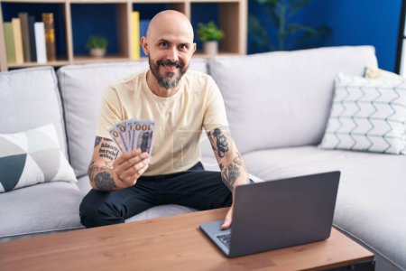 Téléchargez les photos : Jeune homme chauve utilisant ordinateur portable tenant des dollars à la maison - en image libre de droit