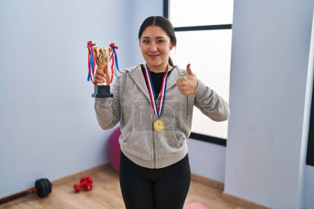 Photo for Young latin woman holding winner trophy smiling happy and positive, thumb up doing excellent and approval sign - Royalty Free Image
